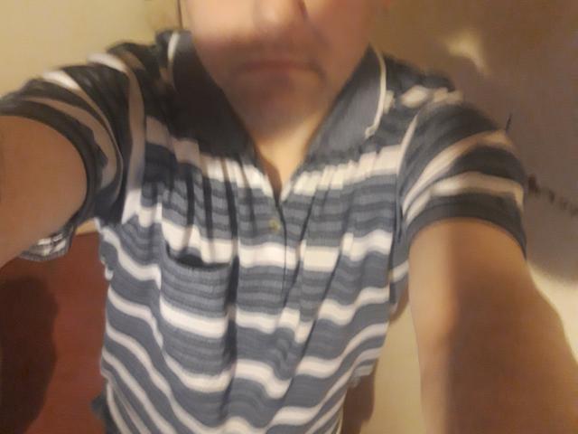 SexyStuff69 in a striped polo shirt, blurred face, slightly open collar, short sleeves, tan skin, and dark hair, with a shadowy background. Focused on upper body.