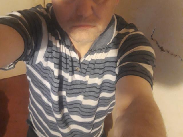 SexyStuff69 with a slightly annoyed face, wearing a striped polo shirt, showing a partial selfie with arms extended. His shirt has dark and white horizontal stripes.