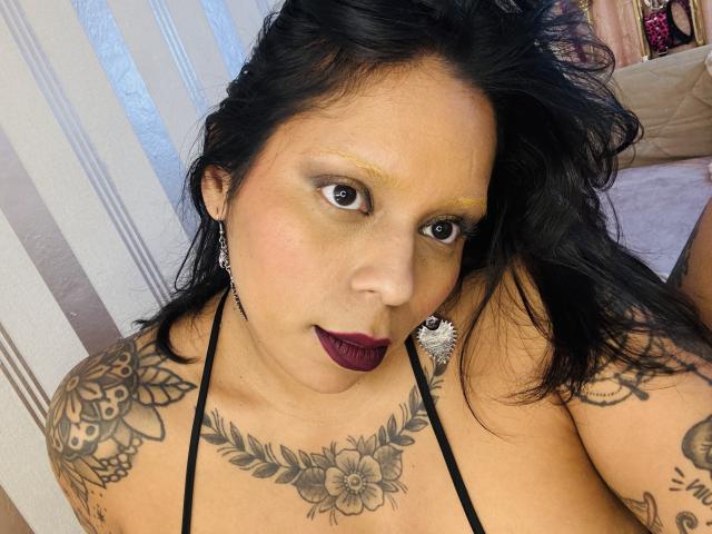 Chili, a sexy Latina slut with dark maroon lipstick and tattoos, gazes at the camera, her black hair cascading.