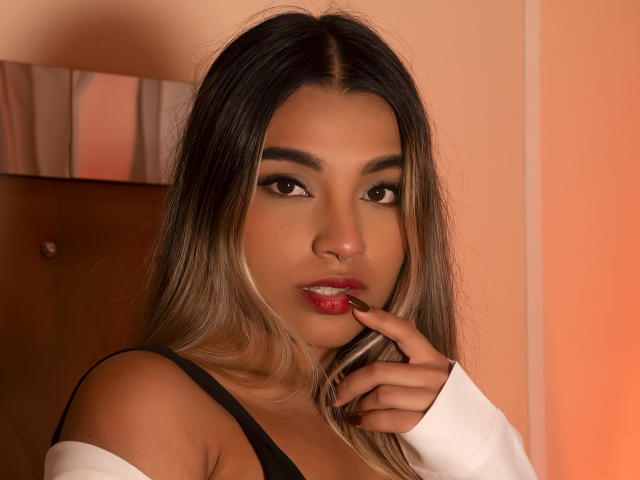 AnneBull with long, dark blonde hair, brown eyes, and red lipstick, finger near her glossy lips, wearing a black strap top and white off-shoulder shirt.