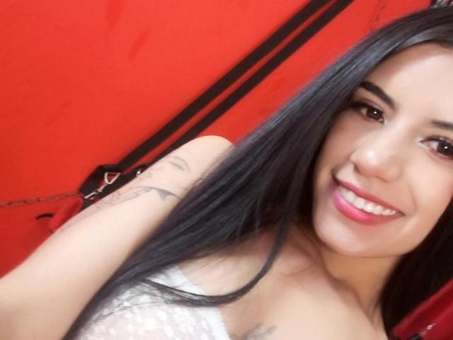 Sami, com cabelo preto longo, batom rosa, sorrindo, blusa branca de renda, tatuagens no braço direito, fundo vermelho, olhando para a câmera, selfie com um leve ângulo..