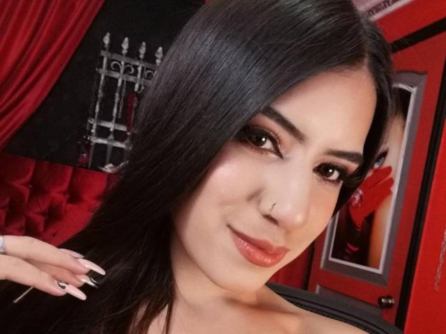 SamiLure tiene el pelo largo y negro, ojos marrones y labios rosados y brillantes. Está sonriendo, lleva un piercing en la nariz, ropa negra y tiene las uñas cuidadas. En el fondo, hay cortinas rojas..