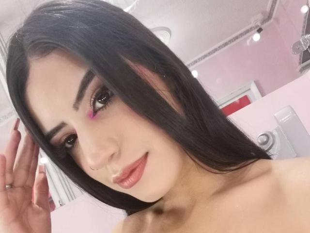 Sami, con lunghi capelli neri, trucco rosa sugli occhi e un piercing al naso, sorride leggermente, con una mano alzata, mostrando le sue spalle scoperte e le labbra rosate..