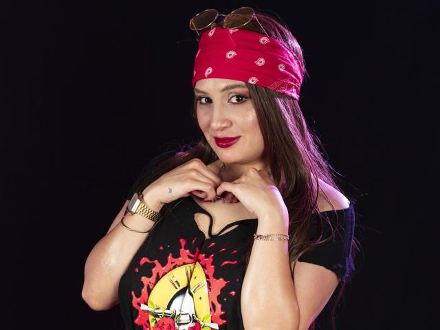 IsabelSoler with long brown hair, red bandana, black off-shoulder tee, sunglasses on head, gold wristwatch, red lipstick, black background, smiling, hands near chest, looking at camera.