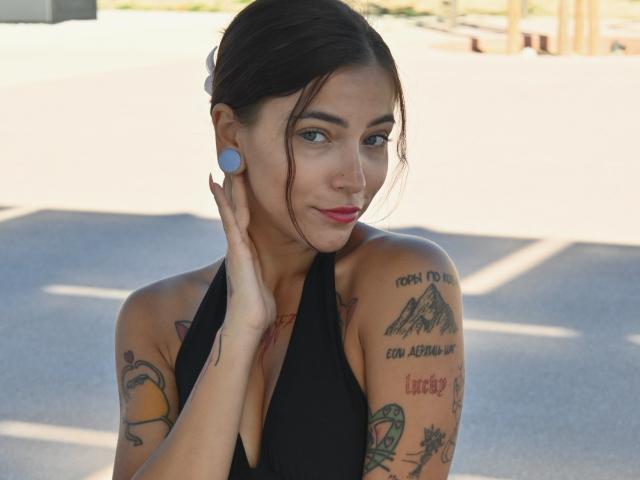 LianiLoverX has dark hair, blue ear plugs, black halter top, tattoos on arms, red lipstick, and touches her cheek with a hand, smiling slightly. Sunlit, outdoor setting.