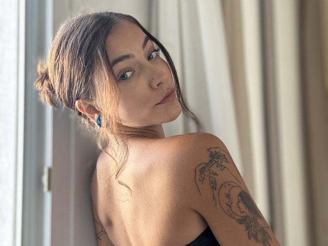 LianiLoverX with brown hair in a messy bun, looking over her shoulder, showing tattoos on her right arm, wearing a black strap top, standing near a window.