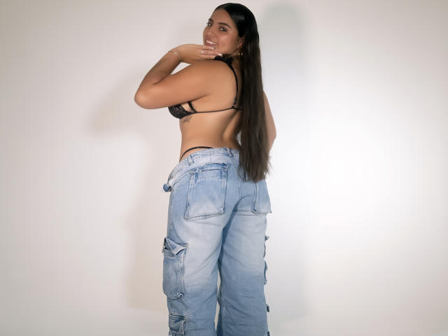 Victoria Rosel mit langen, braunen Haaren, einem schwarzen BH und blauen, weiten, zerrissenen Jeans, die ihren Rücken zeigt und über die Schulter lächelnd, mit der Hand in der Nähe ihres Mundes. Brüste, Hintern..