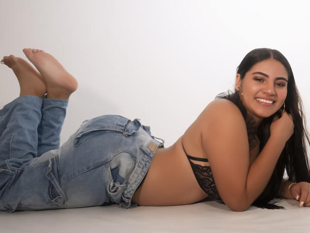 Victoria Rosel, sdraiata a pancia in giù, sorridente, con lunghi capelli scuri, indossa un reggiseno di pizzo nero e jeans blu, mostrando il sedere scoperto e i piedi sollevati in aria..