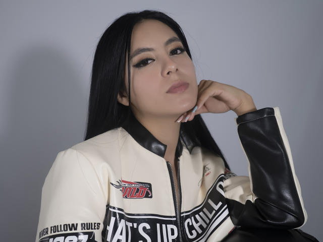 EimmyPark with long black hair, wearing a white and black racing jacket, has a serious expression, resting her chin on her hand, looking slightly to the side.