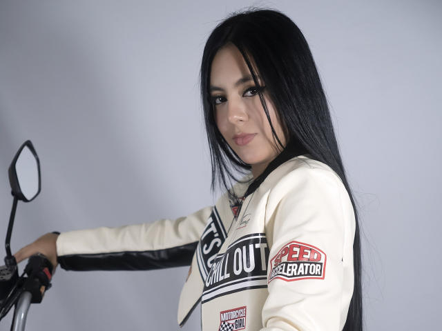 EimmyPark with long black hair, wearing a white racing suit, holding a motorcycle, smiling subtly, right arm raised, left hand on handle, looking at camera.