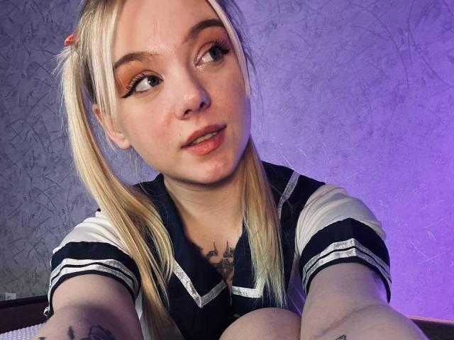 EnchantingEyes with blonde pigtails, wearing a navy and white schoolgirl outfit, has small tits, a tattoo on her chest, and a slightly open mouth, looking to the side.
