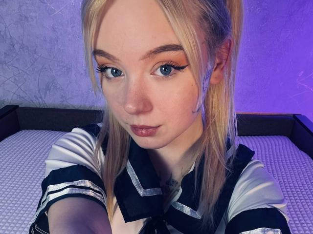 EnchantingEyes with blonde hair, blue eyes, and winged eyeliner, wearing a black and white schoolgirl outfit, has a cute, serious expression and a small nose.