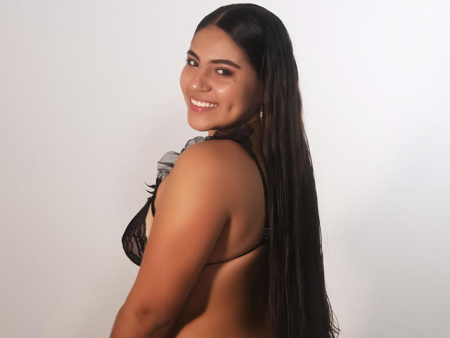 VictoriaRosel, a young woman with long, straight black hair, stands sideways, looking over her shoulder with a smile. She has a medium brown skin tone and is wearing a black lace bra. Her right shoulder is slightly raised, revealing a small, round nose ring on her right nostril. She has a curvy, attractive physique with a visible side profile of her breast. The image has a simple, white background, focusing all attention on VictoriaRosel. Her gold hoop earrings are visible. The lighting is even, highlighting her smooth skin and natural beauty.