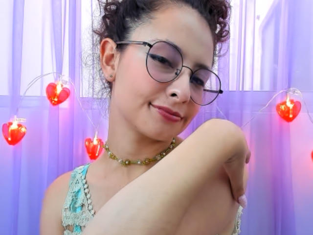 Emma2202 with curly hair, glasses, and pink lipstick, smiling gently, holding her arm, wearing a green lace top and a green beaded necklace, red heart lights in background.