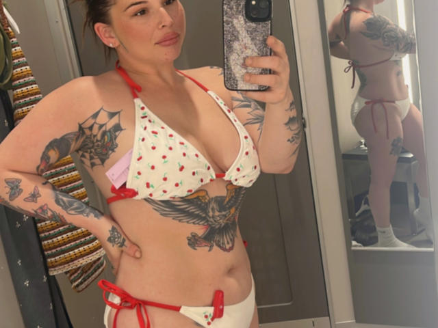 MonyBelle, a tattooed woman with white strawberry-print bikini, shows off her small tits and ass in a mirror selfie, holding a phone with a glittery case.