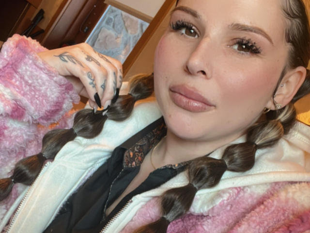 MonyBelle has puffy pink and white fluffy jacket, black lace top, braided brown hair, tattoos on hand, glossy pink lips, and is looking at camera with a slight smile.