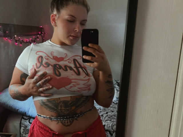 MonyBelle with short brown hair, tattoos on arms and belly, white crop top, red pants, holding phone, covering small tits with hand, mirror selfie, bedroom, pink string lights.