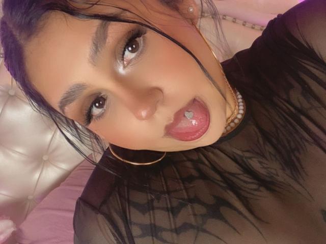 EmmaMyles with dark hair, brown eyes, and pink tongue out, wearing a black sheer top and pearl necklace, has a shiny, wet tongue and hoop earrings.