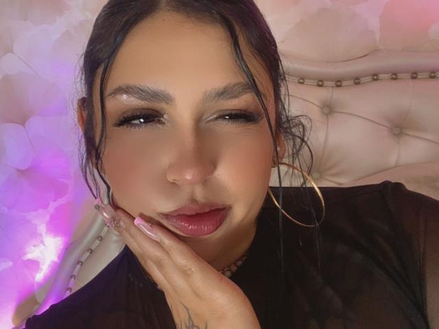 EmmaMyles with dark hair, hooped earrings, and pink lipstick, touching her cheek with pink nails, looking at the camera with a slight smile. She has a nose ring.