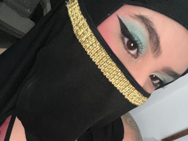 DaliilaAli's face with black mask, gold trim, and blue-green eyeshadow, thick black eyeliner, brown eyes, and a black hijab covering her nose and mouth. Visible tattoo on right arm.