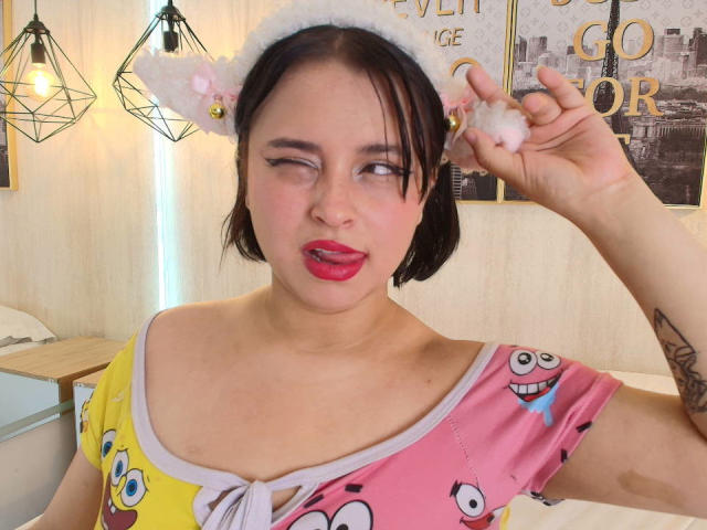 Blueeskyy with short black hair, white fluffy cat ears, and pink nose, winking, tongue out, wearing colorful cartoon print dress, holding fluffy white ball, tattoo on right arm.