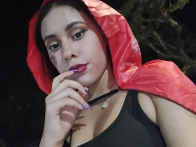 MarilynHills with dark hair, red hood, black tank top, purple lipstick, and purple nail polish, touching her chin, showing her tits, wearing a black choker. Nighttime.