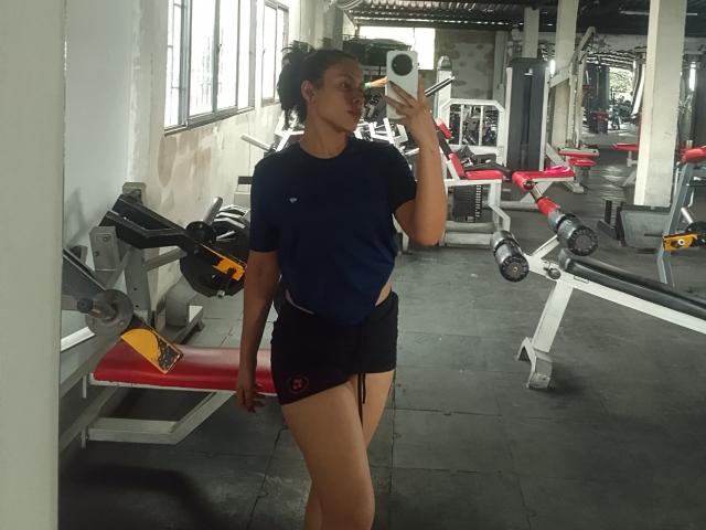MarilynHills, with curly black hair, wearing a blue shirt and black shorts, taking a mirror selfie in a gym, showing her toned ass and small tits.
