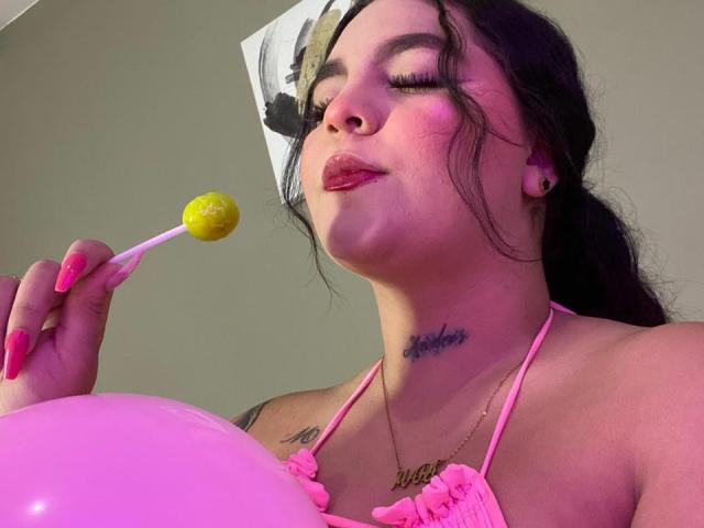 MelodyBlossom con le unghie rosa, che tiene un lecca-lecca giallo, indossa un reggiseno rosa, ha un tatuaggio sotto il mento, tiene un palloncino rosa, ha il rossetto rosa, lunghi capelli scuri e un orecchino, occhi chiusi, labbra strette..