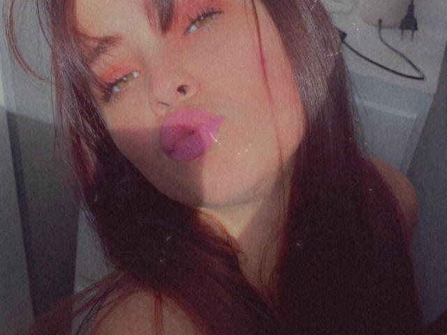 Bellaxx69 with pink lipstick, pouty lips, and half-closed eyes, has long brown hair, fair skin, and a slight shadow on her face. She's making a kissy face.