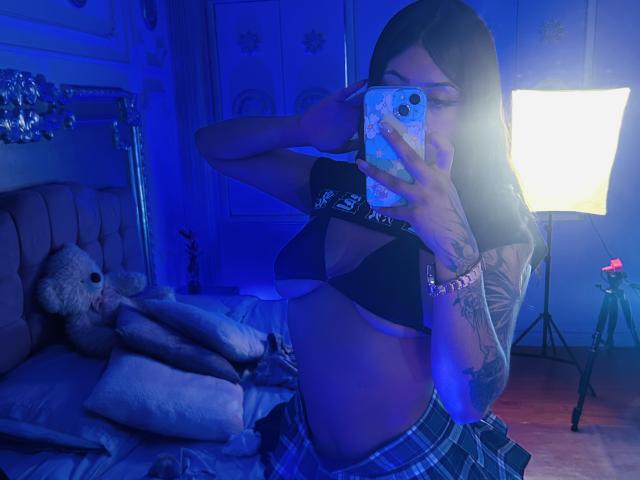 KatianPierce with tattoos, wearing a black bra and plaid skirt, holding a phone in a blue-lit room, mirror selfie showing her tits and a soft toy on the bed.