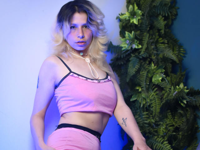 SarahWelinton with blonde, wavy hair wearing a pink, crop-top, revealing her tits, and matching pink shorts, has a tattoo on her right arm, standing near green foliage.