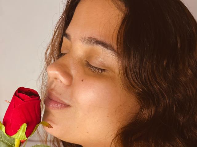 LuisaVanegas with eyes closed, brown wavy hair, and a small mole on her right cheek, gently kissing a red rose. Her lips are slightly parted, showing a soft smile.
