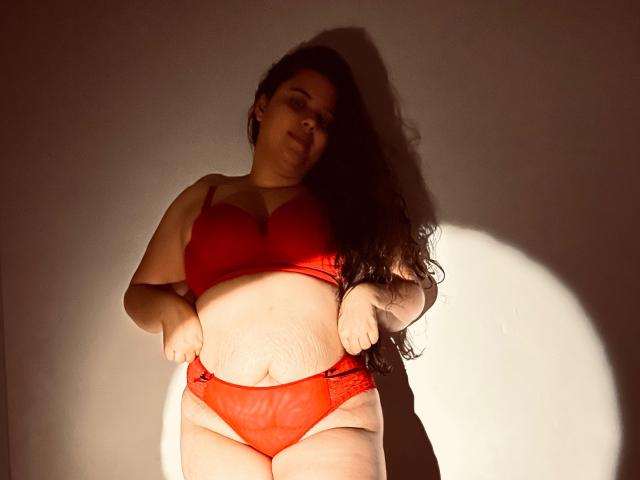 LuisaVanegas with long dark hair, wearing red bra and panties, standing, slightly leaning, hands on waist, soft shadow, light highlighting her belly and red lingerie.
