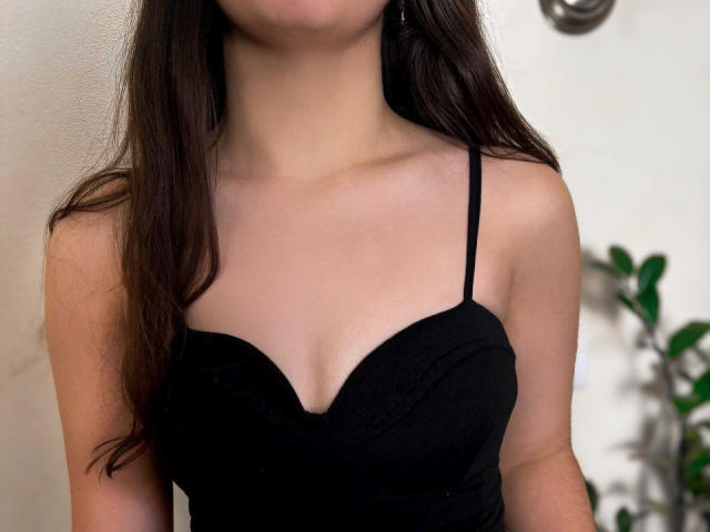 Symphoniia's chest with small tits in a black, strapless, sweetheart-style top. Her brown hair is down, and she's against a white wall with a green plant.