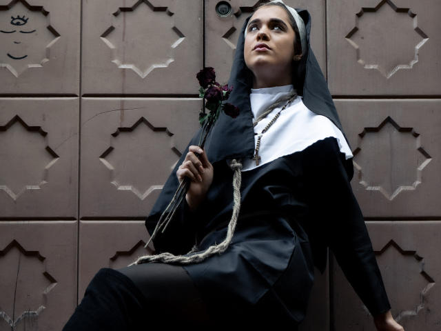 AliceCoolenk, wearing a black and white nun outfit, holds a rose in her hand, sitting against a brown, patterned wall, with a rope tied to her chest.