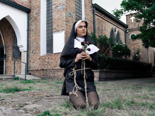 AliceCoolenk, dressed as a nun, kneels on grass with rope tied to her tits, holding rosary beads, in front of a brick church. She has a white wimple and black habit.