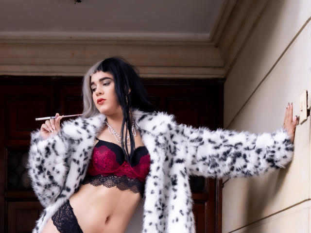 AliceCoolenk stands in a doorway, wearing a white and black spotted fur coat, red lace bra, and black lace panties. She has dark and white streaked hair, bold red lipstick, and holds a white cigarette holder in her right hand. Her left arm is outstretched against the wall. She has a light skin tone, sharp features, and a confident expression. The door behind her is wooden. The image has a sharp, high-contrast look.