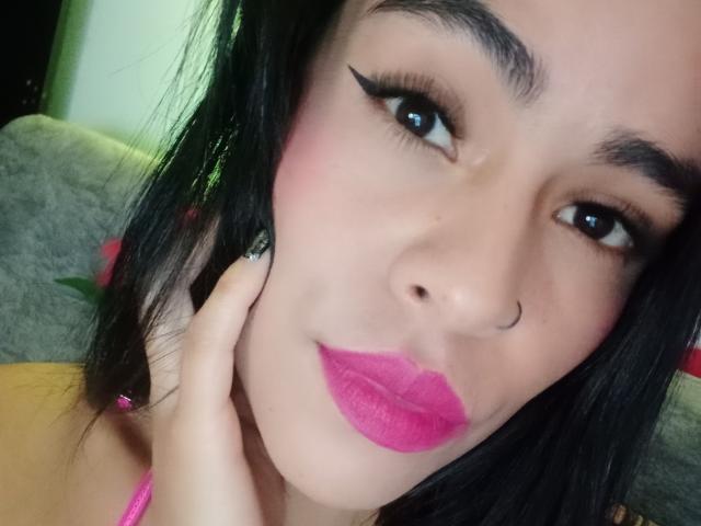 StellaBeckker's face with pink lipstick, black eyeliner, nose ring, and black hair, touching her cheek with her hand, has pink nails and a cute, sexy look.