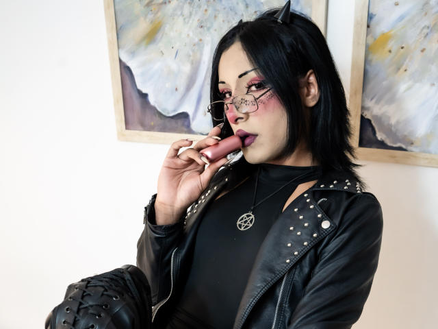NoemiSky, con i capelli neri, orecchie a forma di gatto, occhiali, ombretto rosa, giacca di pelle nera, mentre succhia un sex toy. Indossa una collana con un pentagramma e ha degli stivali neri..