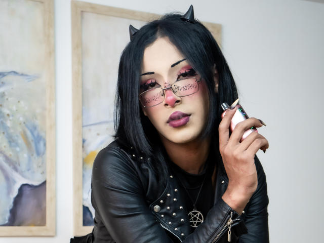 NoemiSky has black hair, wearing cat ears, and pink-tinted glasses with black frames. She's in a black leather jacket with silver studs, holding a white phone to her ear. Her pink lipstick is prominent. Her nails are long and black. She has a pentagram necklace. The background includes two abstract paintings. The image is a photograph.