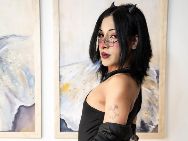 NoemiSky, con i capelli neri, gli occhiali e le corna nere, indossa una canotta nera senza maniche, mostrando un tatuaggio sul braccio destro, ed è in piedi di fronte a dipinti astratti..