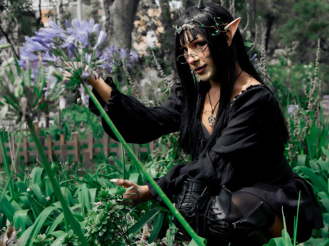 NoemiSky, a cosplay character with elf-like ears, kneels in a lush garden. She has long black hair, wears glasses, and a black off-shoulder top with a deep V-neck. She's touching a large purple flower with green stems. She's wearing black boots with lace details. The greenery and blurred background create a natural, forest-like setting. The photo has a realistic, detailed style.