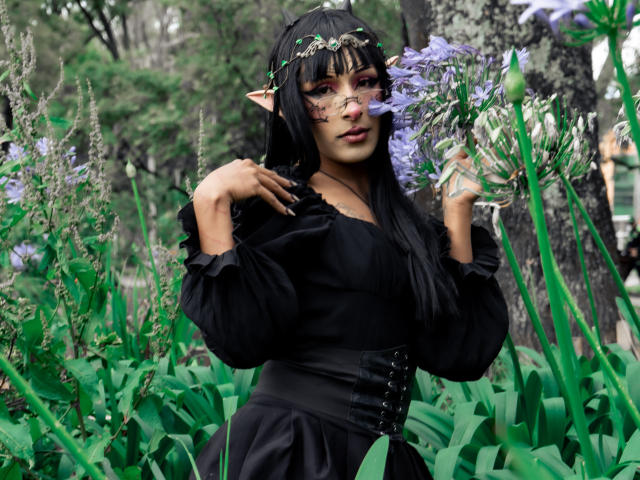 NoemiSky has long black hair, elf-like ears, and wears a leafy headband. She's in a forest, holding lavender flowers, and wears a black dress with a corset. Her makeup is bold with red eyeshadow. She has tan skin and full lips. The green leaves and tall plants surround her. Her right hand touches her hair while her left holds flowers. The photo is natural and vibrant, with a focus on NoemiSky and the floral elements.