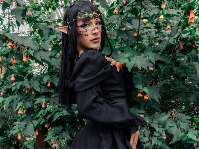 NoemiSky stands in a lush green environment with orange bell-shaped flowers. She has long black hair, elf-like pointed ears, and wears a gold ornate headpiece with pink and red makeup on her face. Her black dress has puffed sleeves and a slightly torn lower section. She looks over her shoulder with a slightly parted lip, her right hand touching her chest. The green leaves and flowers create a dense, natural backdrop. The image uses vibrant colors and contrasts the dark dress with the bright floral background.