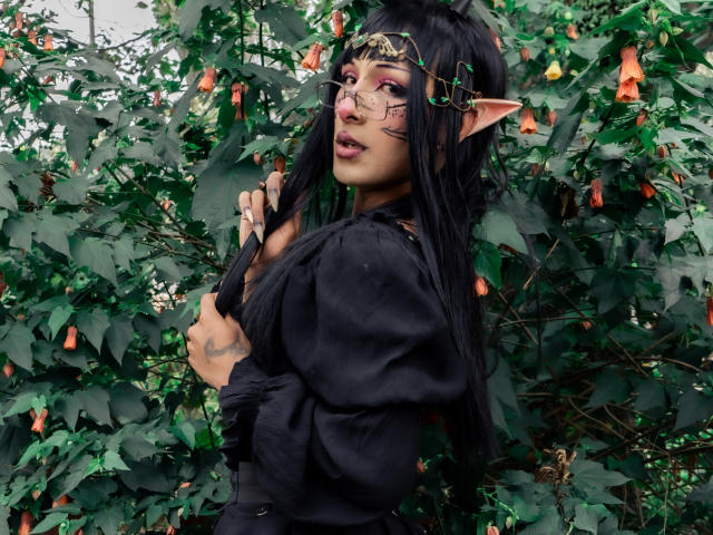 NoemiSky stands in a lush, green environment with leafy plants and orange flowers. She has long black hair, elf-like ears, and wears gold headpiece with green vines. Her makeup includes pink eyeshadow and facial tattoos. She wears a black, puffy-sleeved top and holds her hair with one hand. Her expression is slightly open-mouthed, with a subtle, confident look. The image is vivid, with natural greenery contrasting her dark attire.