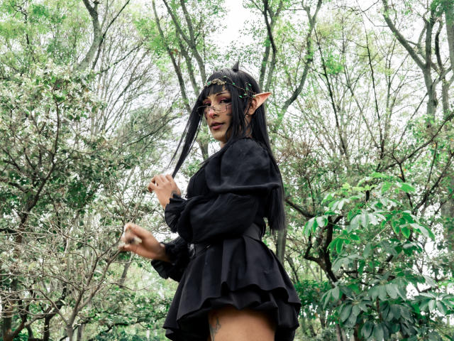 NoemiSky stands in a forest with tall trees. She has long black hair, pointed elf ears, and dark makeup. She wears a black, puffy-sleeve dress with a layered skirt. Her right hand is raised, and she looks over her shoulder with a slight smile. The forest is green and dense with leaves. Her tattoo is visible on her right thigh. The overall style is fantasy cosplay. The composition emphasizes her elf-like features and forest setting.