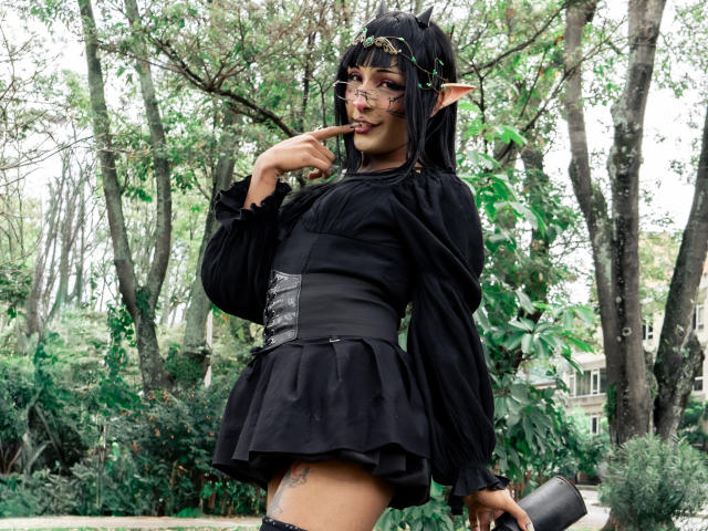 NoemiSky stands in a forested area, wearing a black, long-sleeve, floral-embellished dress with a corset-like waist. She has long black hair, elf-like ears, and is sucking on a finger. Her skin is dark, and she has a tattoo on her right thigh. She holds a black cylindrical object in her right hand. The greenery and trees in the background are blurred. The image is clear with natural light. NoemiSky's expression is playful. The style is cosplay with fantasy elements.