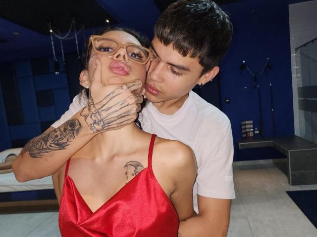 LoveBoundDuo with glasses, red satin dress, tattoos, and pouty lips, has face gently held by LoveBoundDuo in white shirt, short hair, and ear piercing, in a blue room.