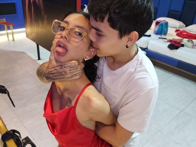 LoveBoundDuo with tattoos gripping LoveBoundDuo's neck, tongue out, in red dress. LoveBoundDuo, in white tee, kisses neck, holding. Visible tattoo on LoveBoundDuo's chest.
