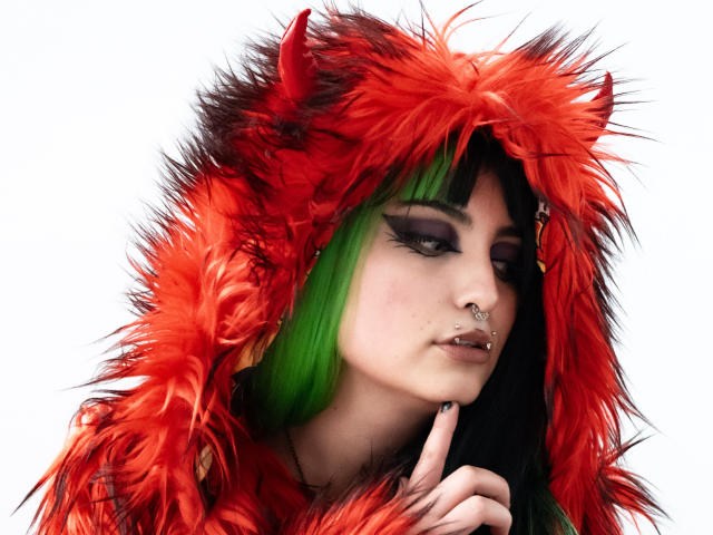 AngelBlakee wears a bright red, fluffy hood with black spikes, contrasting her green hair and dark roots. She has heavy black eyeliner, a nose ring, and black nail polish. Her finger is near her lips in a thoughtful pose. The red hood has a textured, almost furry appearance, while her green hair is sleek and straight. The image has a white background, emphasizing the vivid colors and bold makeup. The overall style is edgy and rebellious.